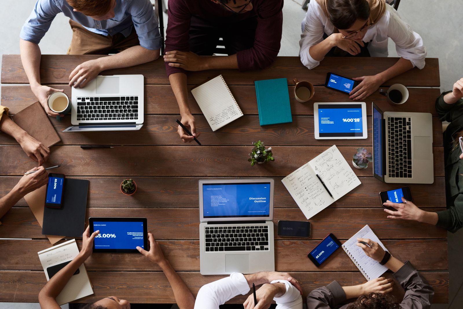 A table seen from above with laptops, notebooks, pens and cups of coffee, and several people working around it.