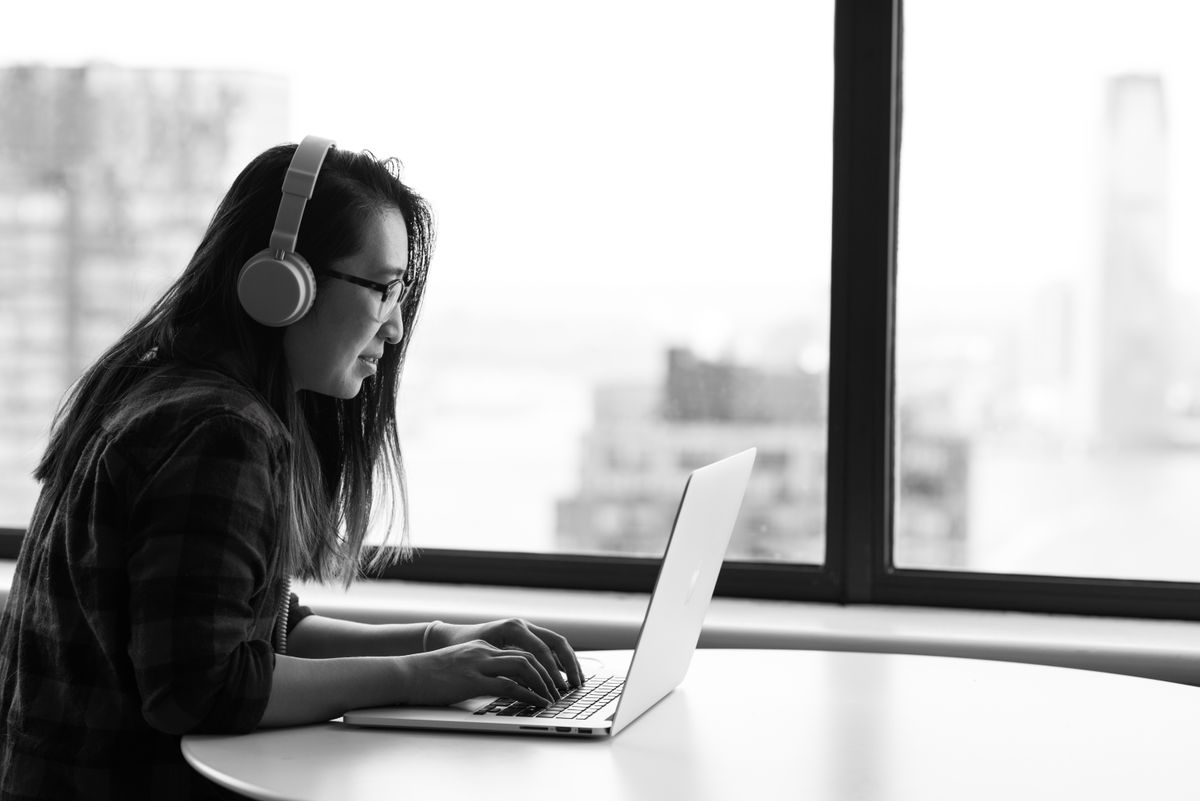 Somebody typing on a laptop by a window in black and white, wearing headphones, a city behind the glass.