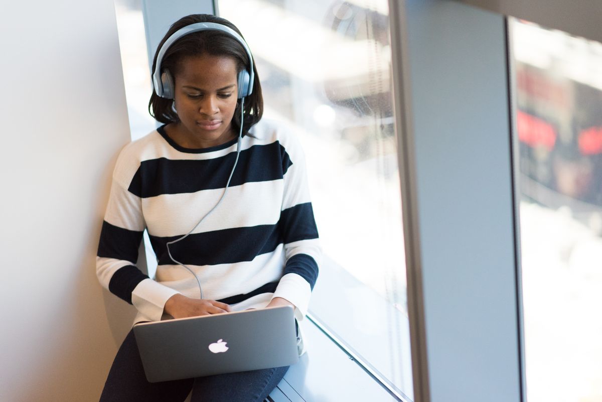 Somebody working on a laptop at a window, wearing headphones, with daylight behind them.