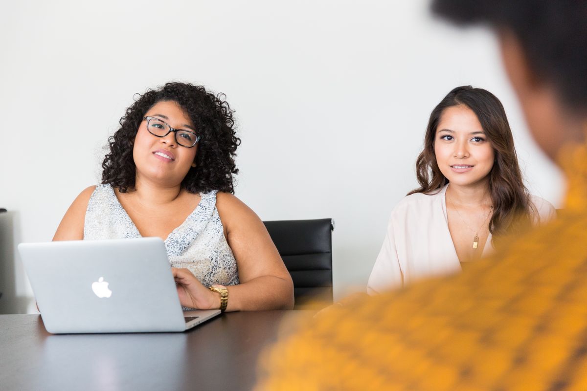 Two colleagues at a table with an open laptop between them, looking up from their work.