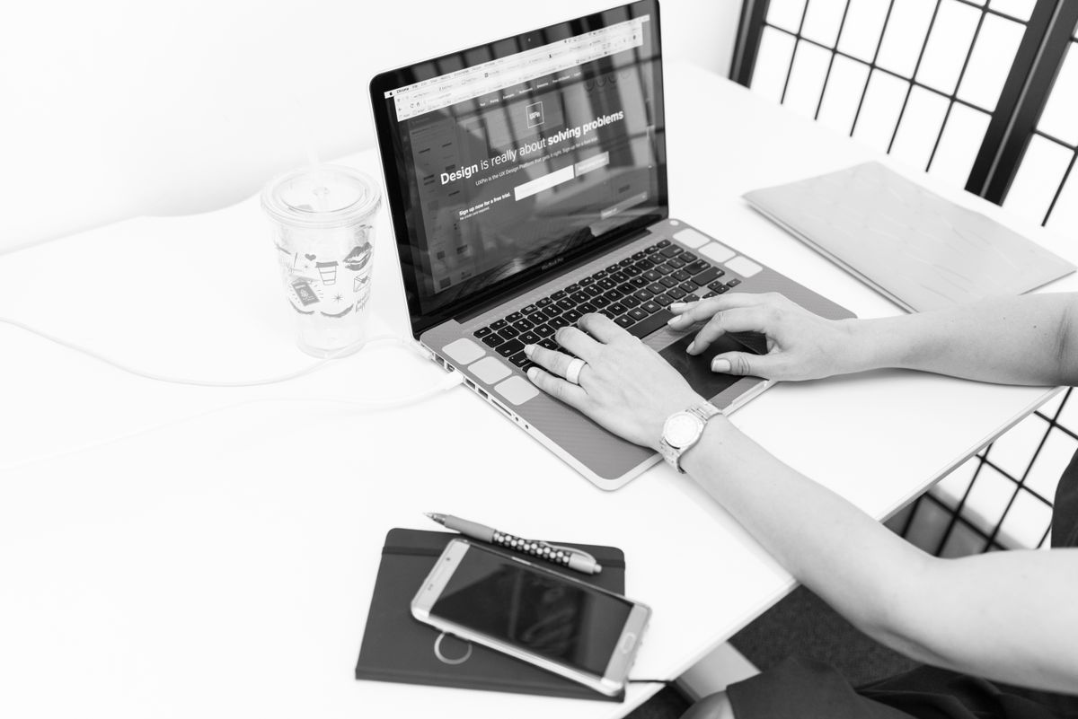 A desk seen from above in black and white: hands on a laptop keyboard, a notebook, a pen, a phone and a coffee cup.